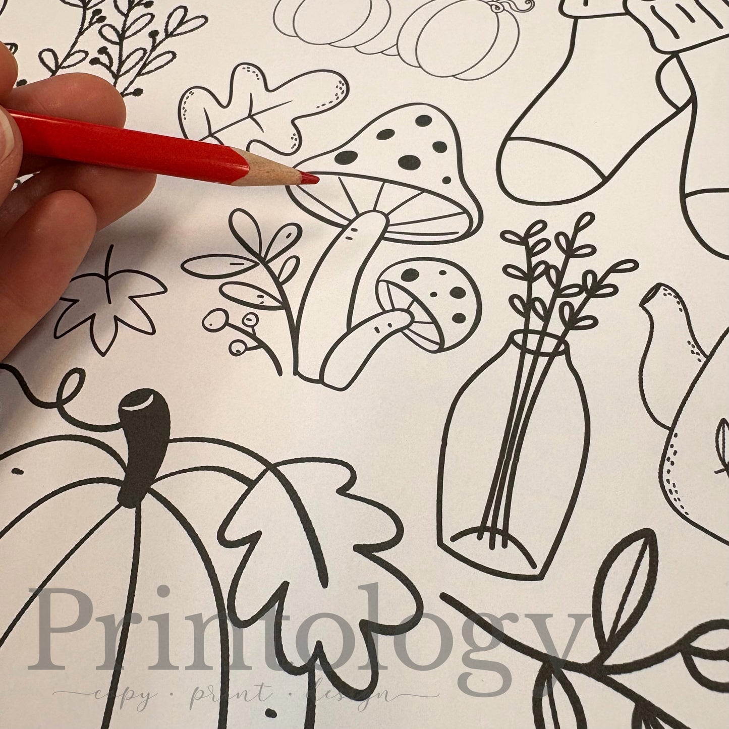 Hand holding a red pencil over a coloring page with black line art of autumn-themed designs including pumpkins, a kettle, mushrooms, fall leaves, and socks.