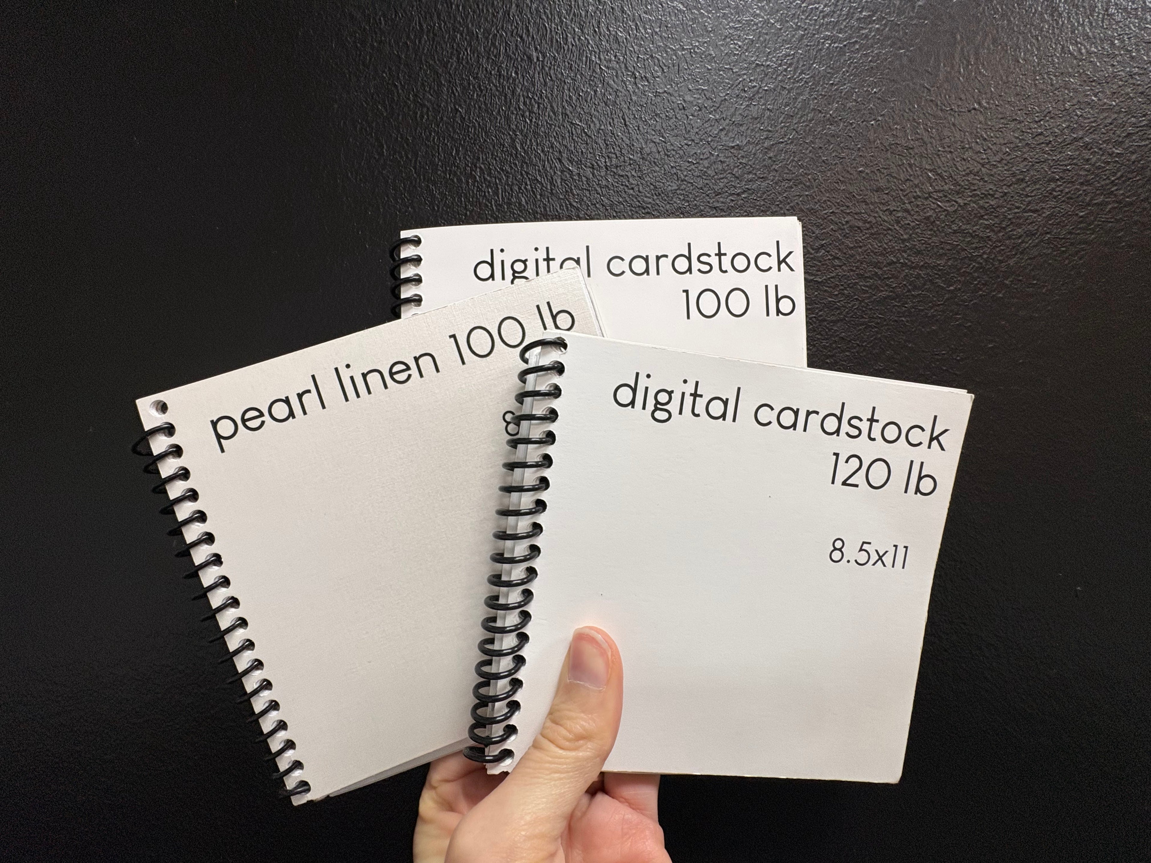 Three spiral-bound digital cardstock samples held against a black background