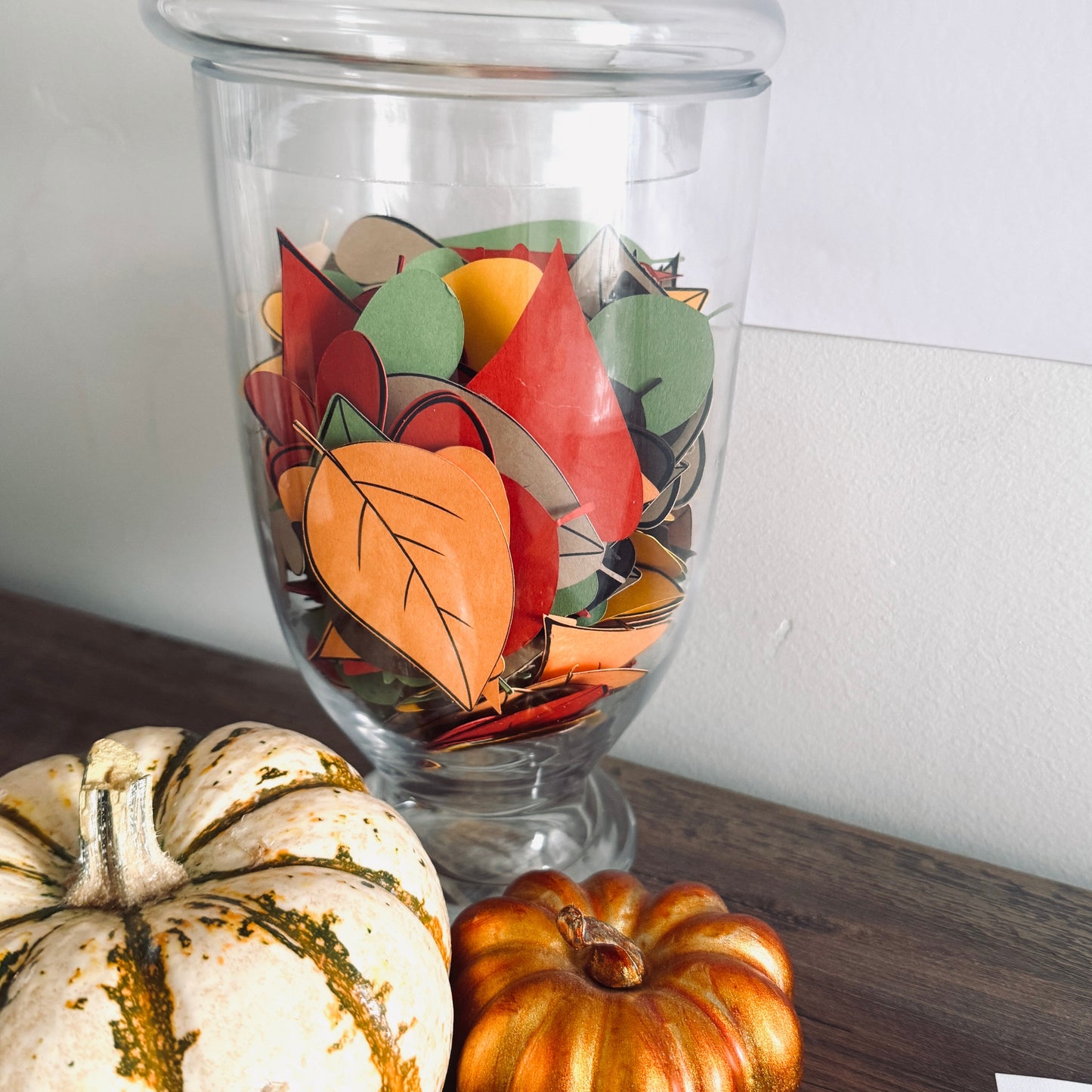 Colorful leaf cutouts in a glass container on a wooden surface with pumpkins.