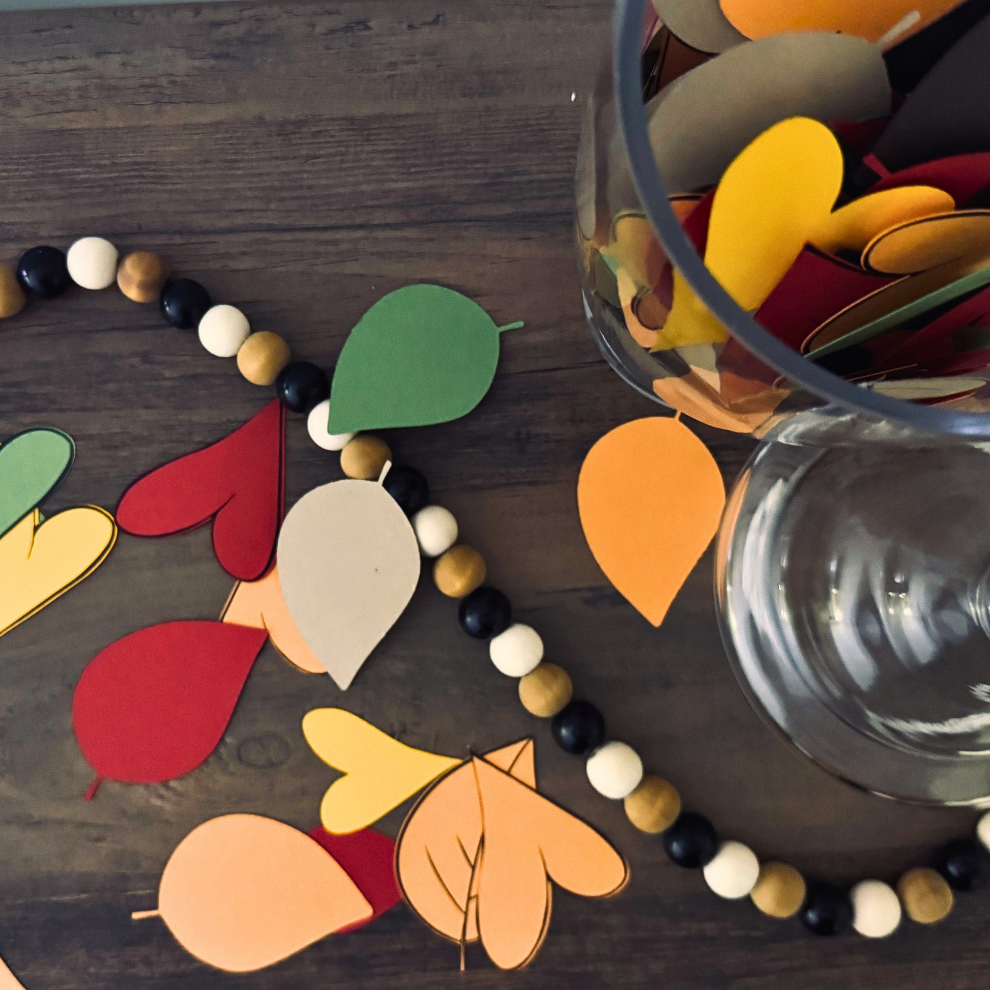 Colorful leaf-shaped cutouts on a wooden surface with a glass container filled with more leaf cutouts.