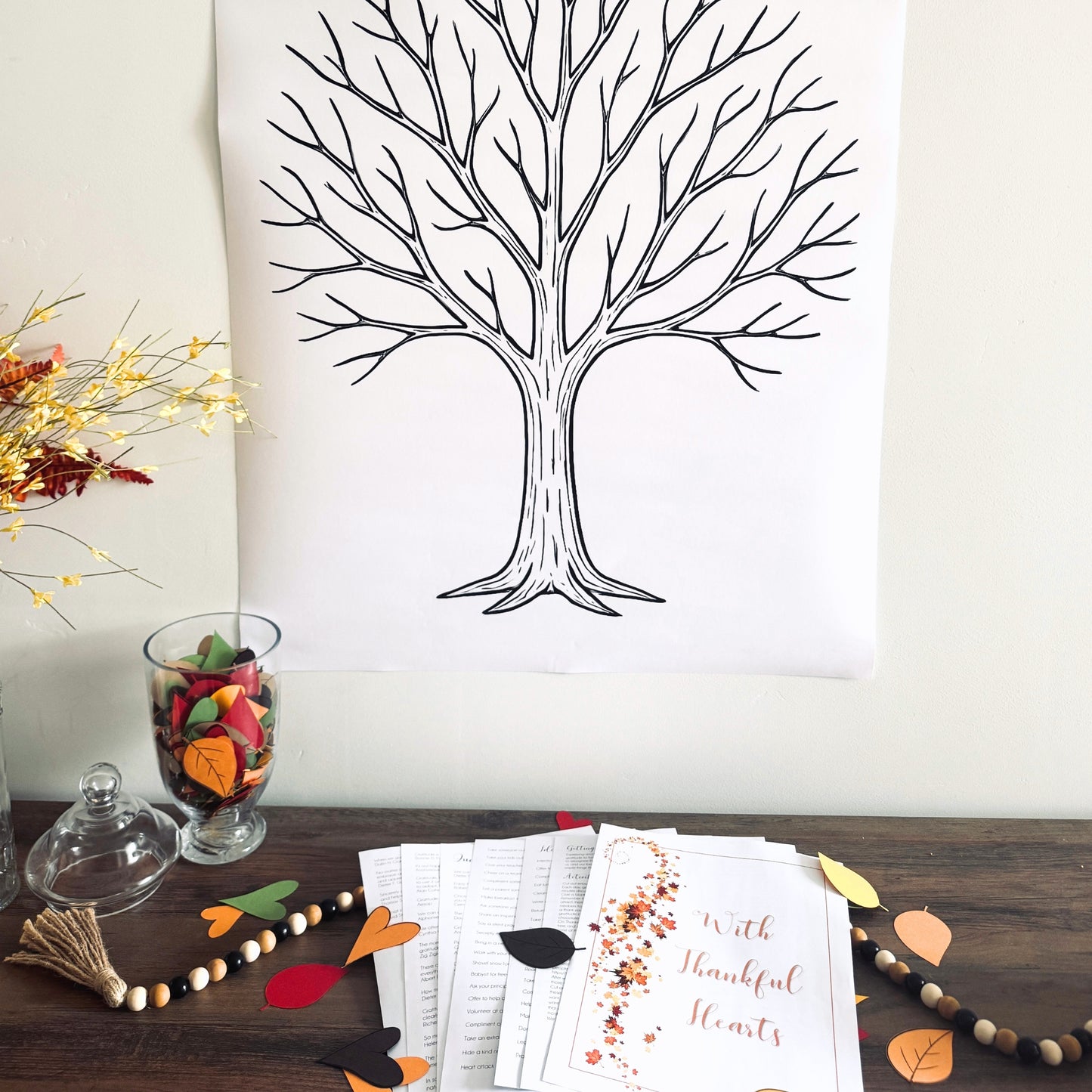 Decorative setup with a tree drawing poster, fall-themed thankful packet, and decorative items on a wooden surface.